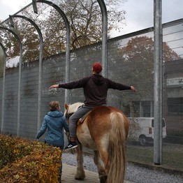 Der Mann reitet auf dem Pferd an einem hohen Sicherheitszaun entlang. Dabei hat er beide Arme ausgestreckt. Die Frau führt das Pferd. Bild: LWL/SchuFi