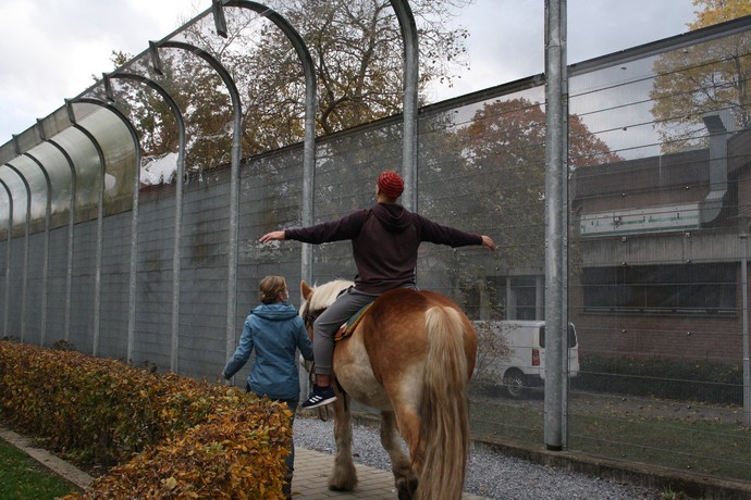 Der Mann reitet auf dem Pferd an einem hohen Sicherheitszaun entlang. Dabei hat er beide Arme ausgestreckt. Die Frau führt das Pferd.