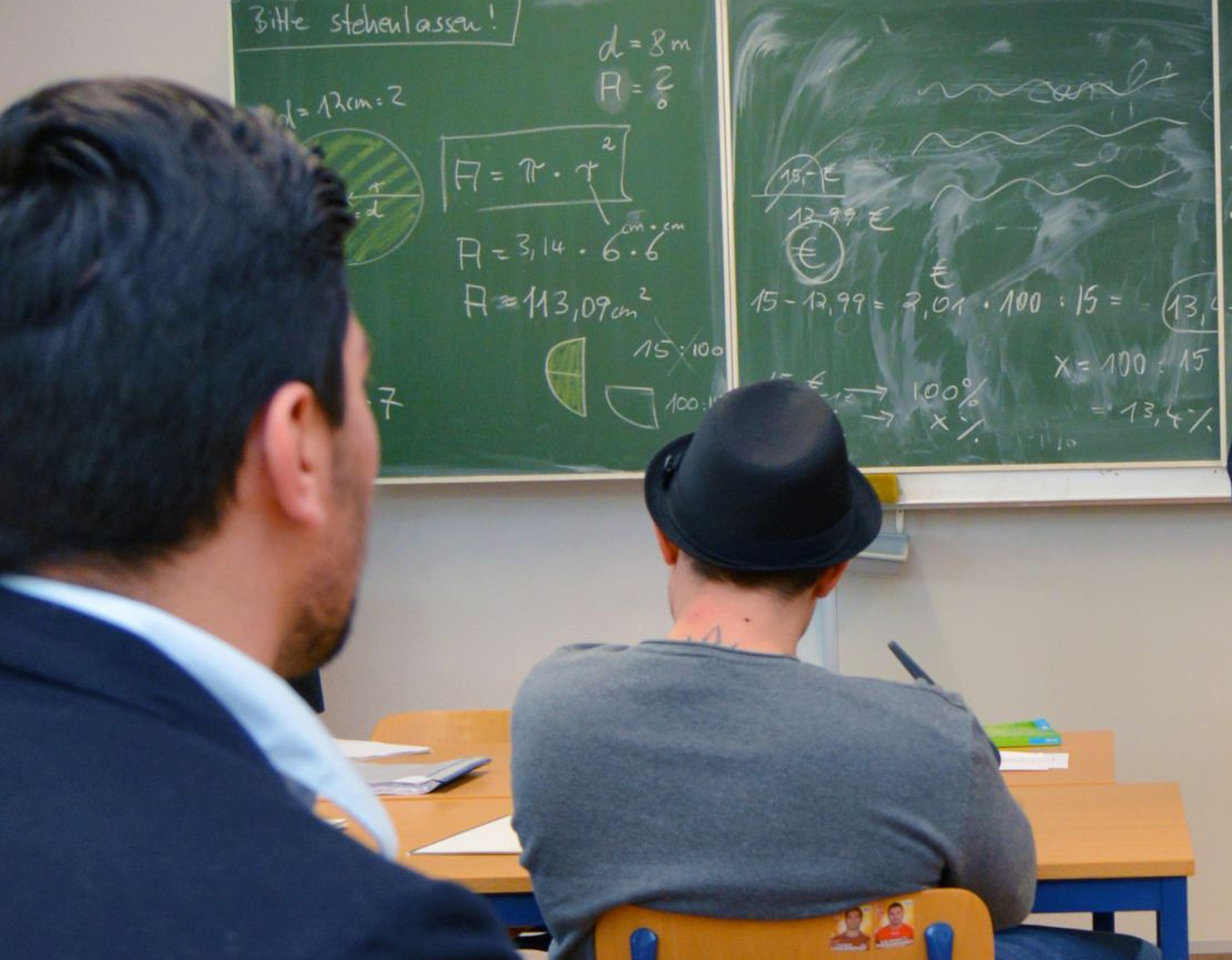 Zwei junge Männer sitzen auf Stühlen an Tischen vor einer mit mathematischen Formeln beschriebenen Tafel. (Bild: LWL/Hannig)