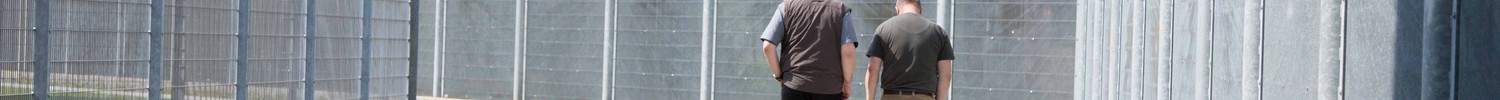 Zwei Männer laufen an einem hohen Zaun entlang. (Bild: LWL)