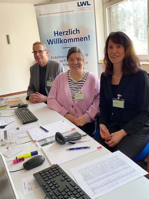 Ein Mann und zwei Frauen sitzen an einem Schreibtisch vor einem Tagungsplakat (Foto: LWL/SchuFi) (vergrößerte Bildansicht wird geöffnet)