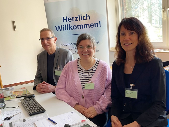 Zwei Frauen und ein Mann sitzen an einem langen Schreibtisch mit Tagungsplakat und lächeln in die Kamera. (Bild: LWL/Schufi)